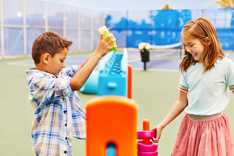 Two children playing with colorful toys on an outdoor tennis court with blue net