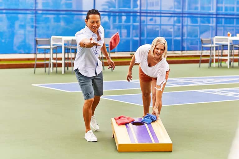 Two people playing cornhole on a tennis court with blue and green courts visible in background