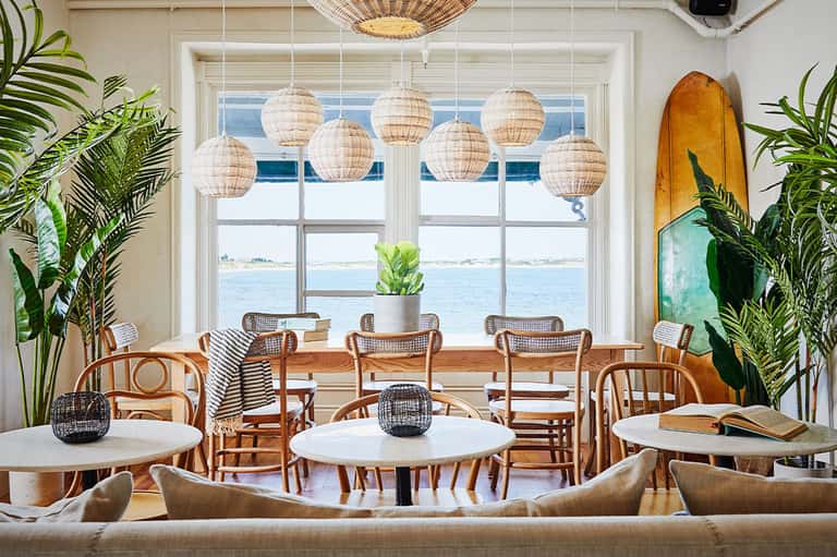Coastal cafe with woven pendant lights, rattan chairs, white tables, ocean view windows, and tropical plants