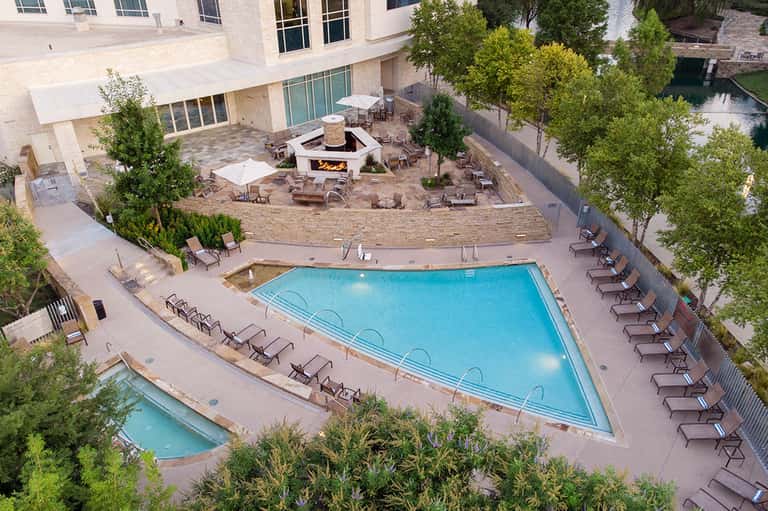 Aerial view of resort pool with lounge chairs, fire pit, and hotel building surrounded by trees