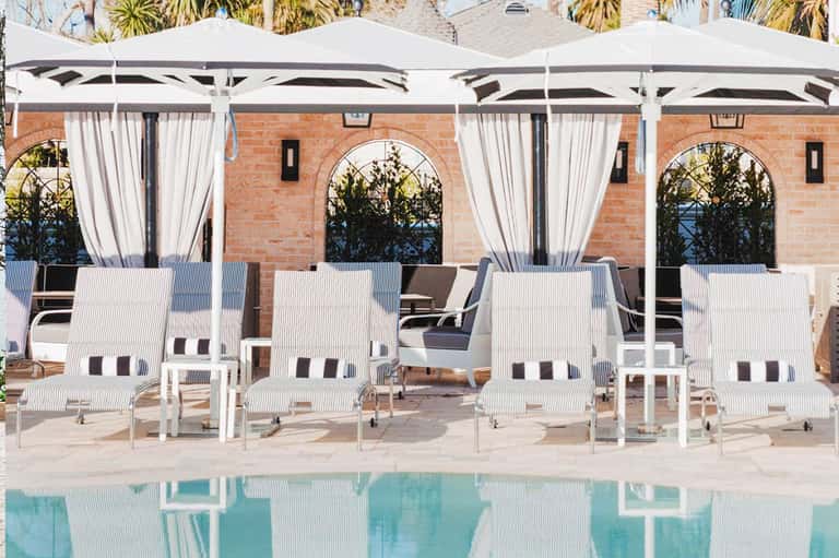 Luxury poolside cabanas with white umbrellas and striped loungers overlooking a turquoise pool against a brick building