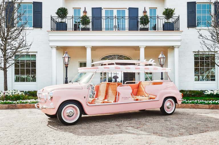 Pink vintage convertible with striped interior parked in front of white colonial-style building with balcony