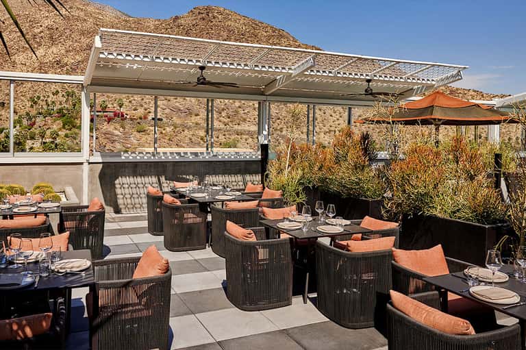 Outdoor dining terrace with dark wicker furniture, orange cushions, and desert mountain backdrop