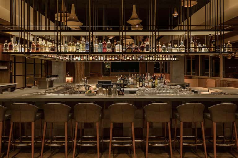 Sophisticated bar with wooden chairs, extensive bottle collection, pendant lights, and modern design