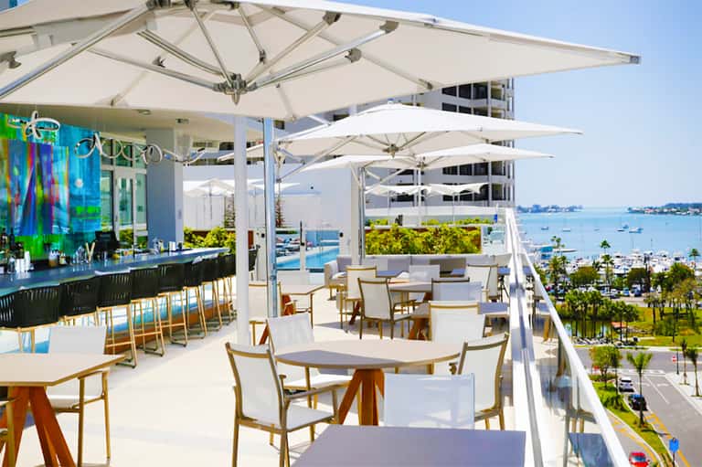 Waterfront restaurant patio with white umbrellas, wooden chairs, bar area, and bay views