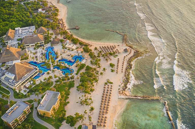 Beachfront aerial view showing resort pools, sandy beach, palm trees, and ocean waves
