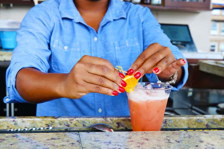 Bartender in blue shirt garnishing a coral-colored cocktail with a yellow citrus twist at the bar