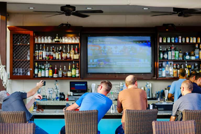 Sports bar interior with patrons watching game on large screen, full liquor shelves, and woven seating