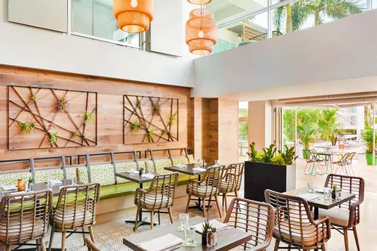 Modern restaurant interior with wooden wall art, woven chairs, and orange pendant lights overlooking outdoor seating area