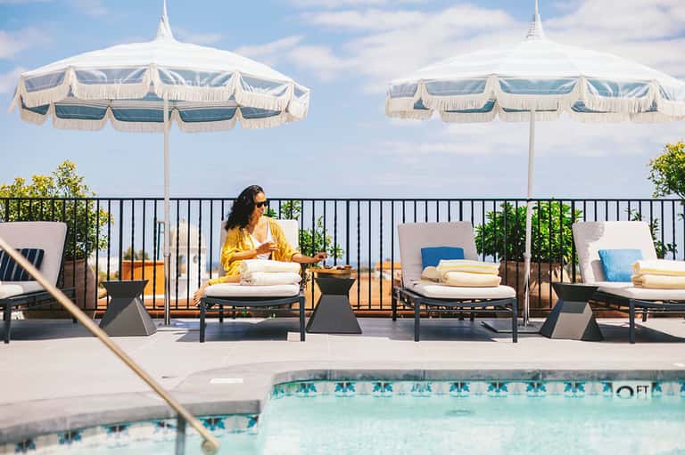 Woman relaxing on pool lounge under decorative umbrella with mountain views beyond railing