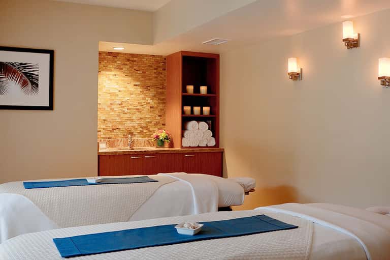 Spa treatment room with massage tables, brick accent wall, wooden shelving with candles and rolled towels