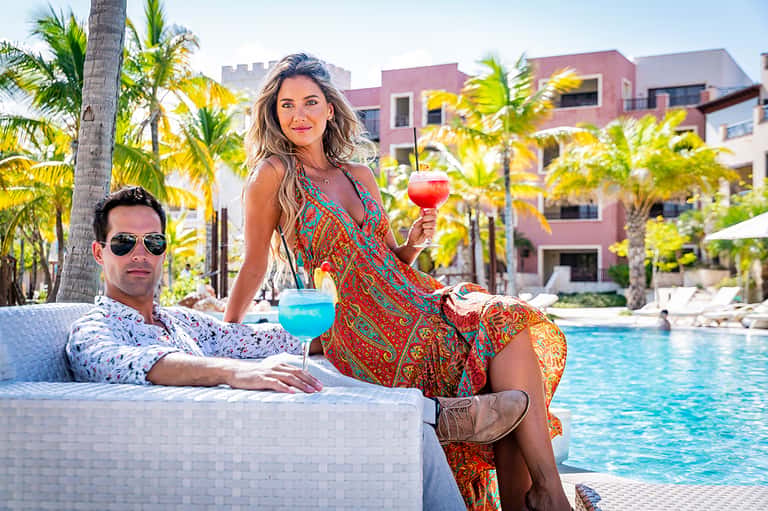 Couple enjoying colorful cocktails in resort infinity pool with pink buildings and palm trees