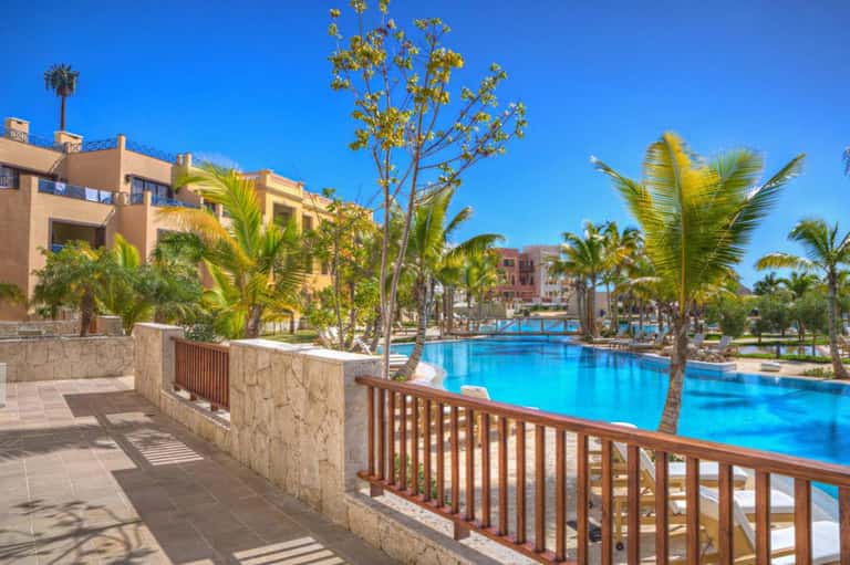 Resort courtyard with sprawling blue pool, palm trees, stone buildings, and sunny day