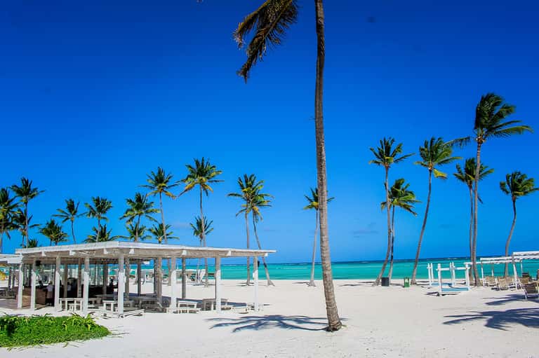 White beach pavilion with turquoise ocean, palm trees, white loungers, and clear blue sky