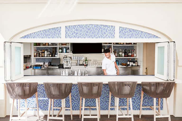 Modern bar with blue tile backsplash, stainless steel counter, and five woven chairs facing bartender in white shirt