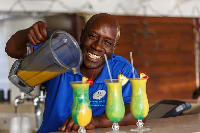 Bartender pouring tropical cocktail, smiling at camera with yellow and green drinks on counter