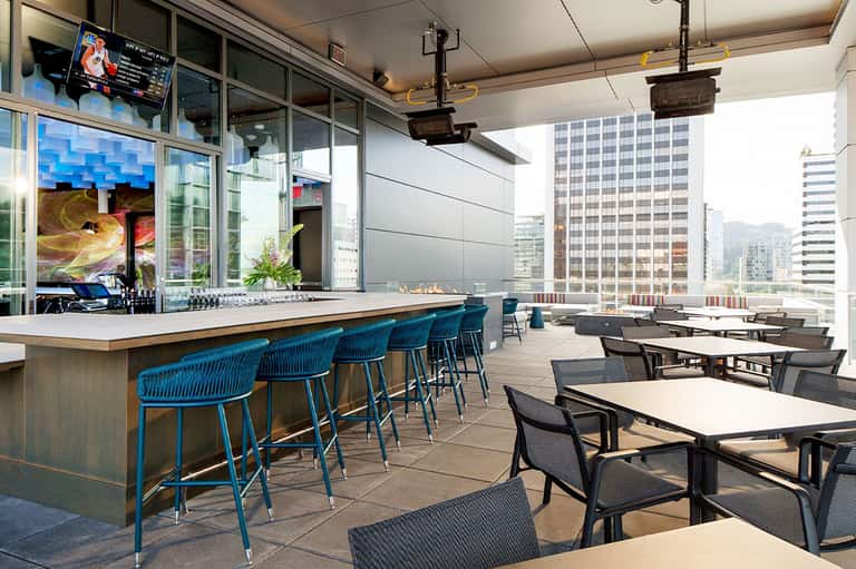 Rooftop bar and lounge with blue bar seating, tables, city skyline views, and floor-to-ceiling windows