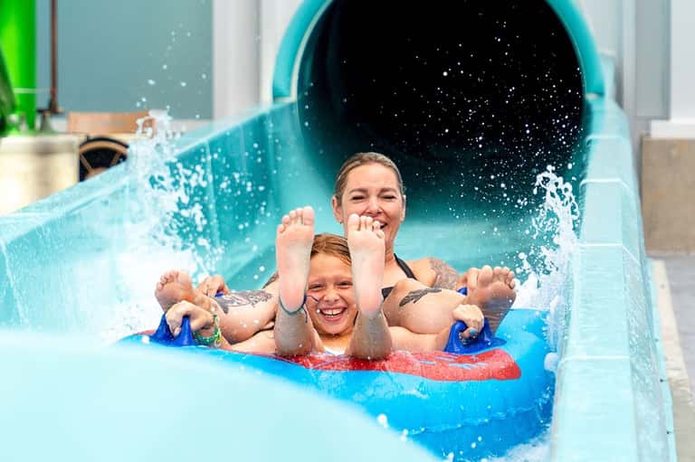 Adult and child on red raft exiting blue water slide with splashing water