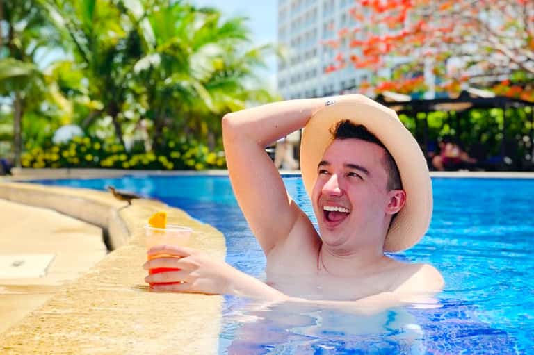 Man in white hat enjoying a resort swimming pool with city buildings and tropical plants in background