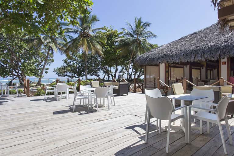 Beachfront terrace with white modern chairs, palm trees, and thatched-roof structures