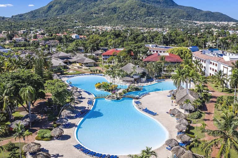 Aerial view of large lagoon-style resort pool complex with blue water, palm trees, and volcano backdrop