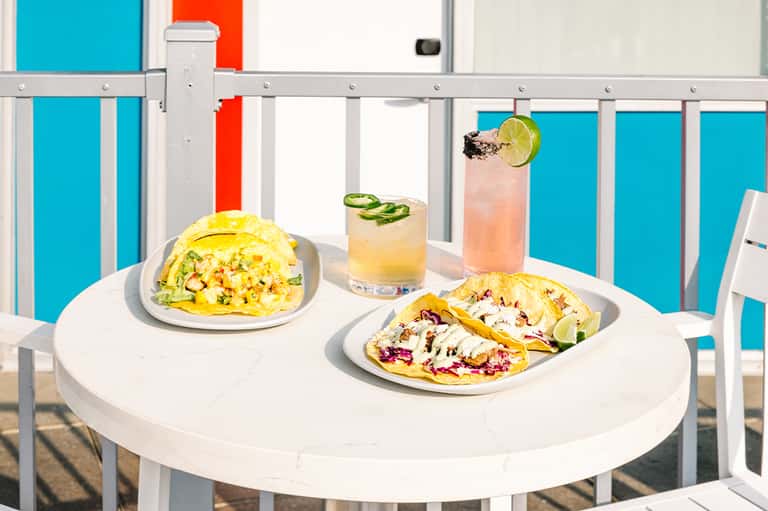Colorful tacos and cocktails on white table with turquoise pool visible behind railing