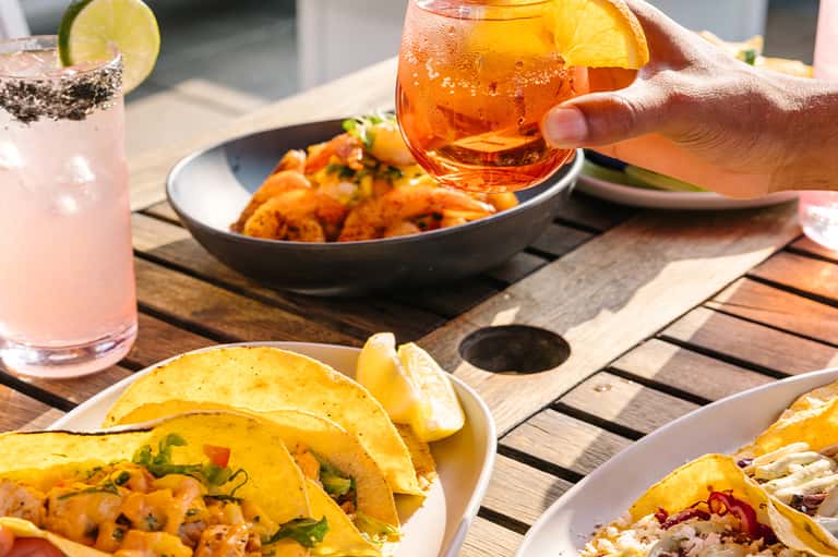Outdoor dining scene with colorful cocktails, tacos, appetizers on wooden table in sunlight