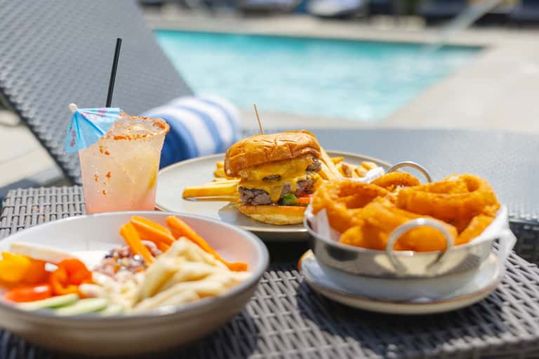 Poolside lunch setting with gourmet burger, onion rings, cocktail, and striped lounge chair