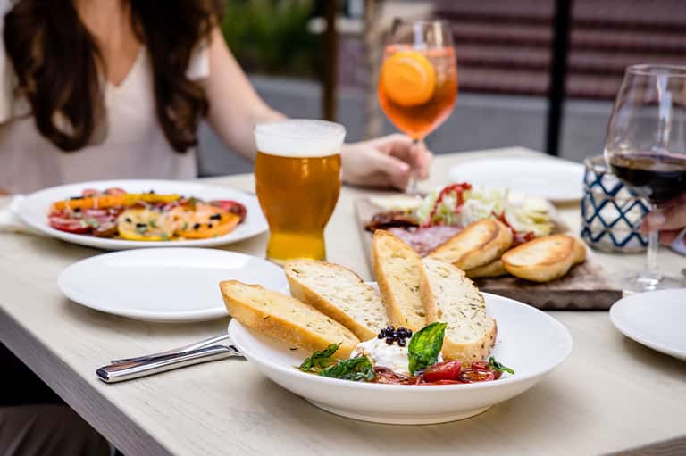 Outdoor dining scene with person holding cocktail, beer, and appetizers on white tablecloth