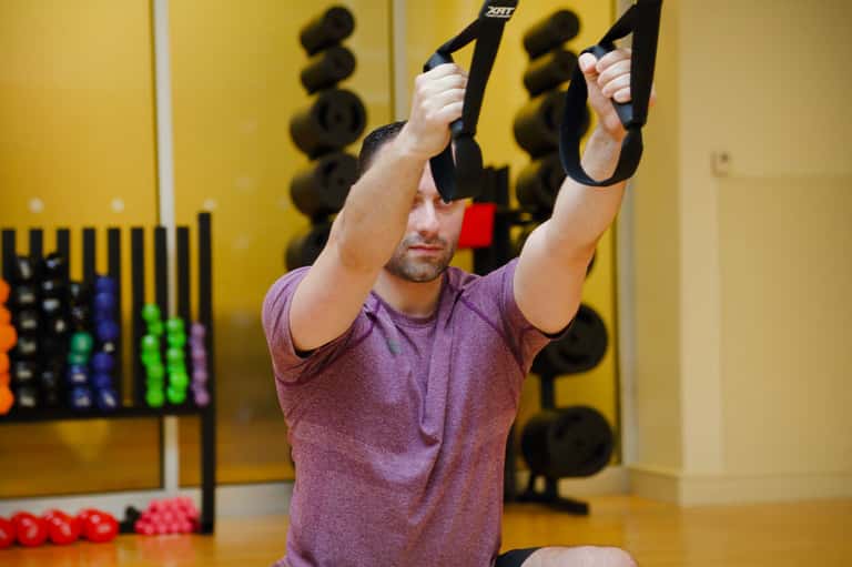 Man in purple shirt performing suspension training exercise with black TRX straps in fitness gym