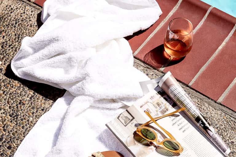 Poolside relaxation scene with white towel, sunglasses, drink, and magazine on deck