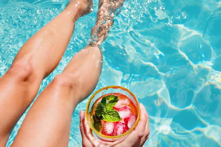 Person holding colorful tropical cocktail by pool with legs in turquoise water