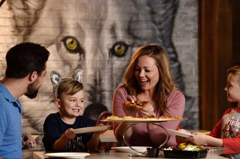 Family dining with playful wolf mural backdrop, adults and children sharing pizza and casual conversation