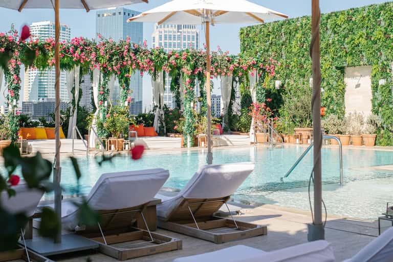 Resort pool with lounge chairs, umbrellas, lush flowering vines, city skyline
