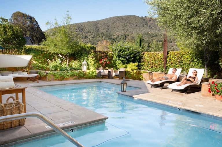 Resort pool area with spa, loungers, green hedges, mountain view, and wooden deck