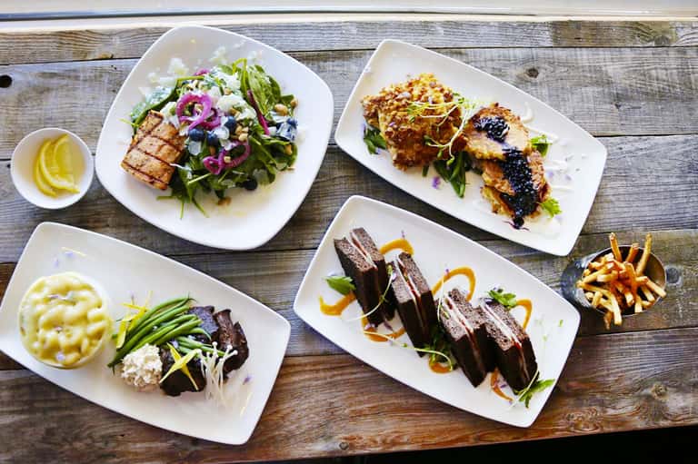 Fine dining plated dishes on wooden table including grilled fish, seared meat, and vegetable sides