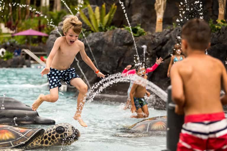 Children playing in resort pool with water fountains and tropical landscaping