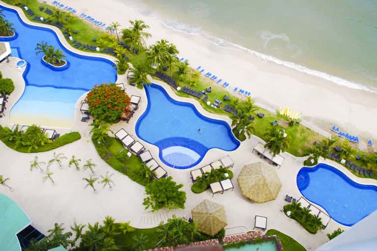 Aerial view of resort pools and beach with palm trees and lounge chairs along sandy shore