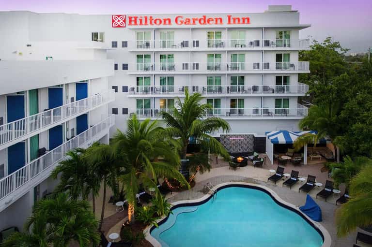 Hilton Garden Inn exterior with white balconies, palm trees, and curved pool at dusk
