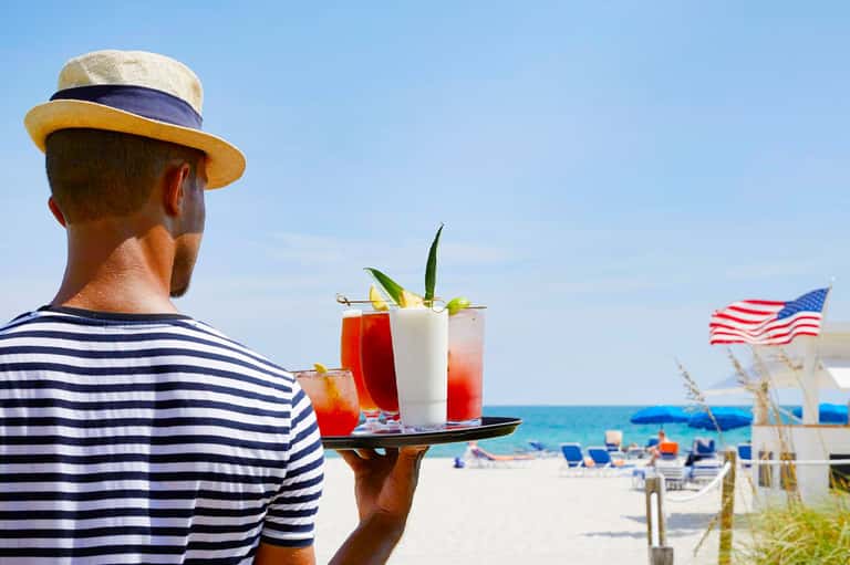 Waiter in striped shirt carrying colorful cocktails on beach with American flag and ocean view