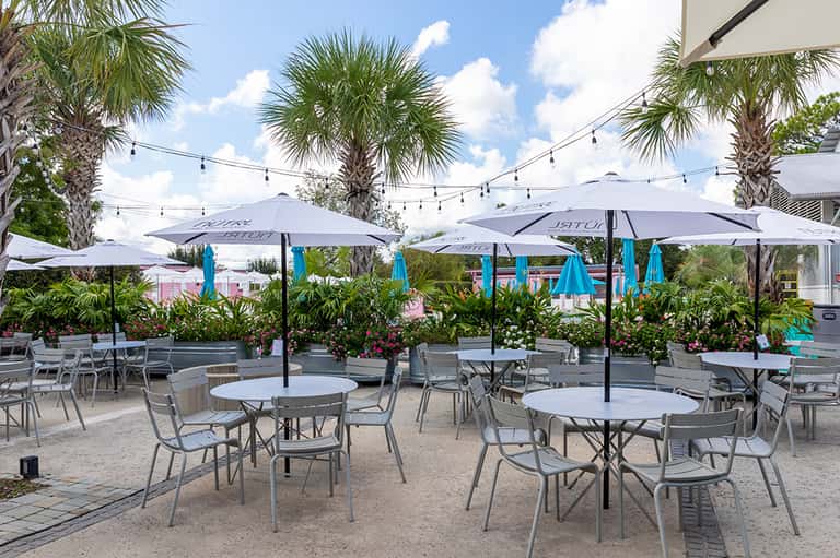 Outdoor patio dining area with white umbrellas, palm trees, string lights, and flowering plants