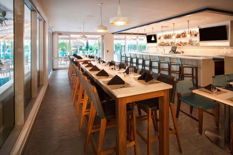 Modern dining room with long wooden table, bar counter, and pool views through glass doors