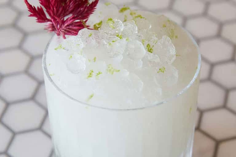 Elegant cocktail with whipped cream, lime zest, and pink flower garnish on hexagonal tile
