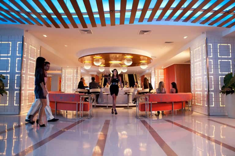Modern hotel lobby with red seating, geometric wall panels, decorative ceiling, and check-in desk