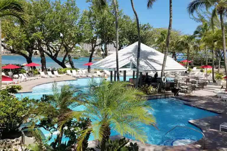 Resort pool area with multiple pools, white tent pavilion, red umbrellas, and waterfront views