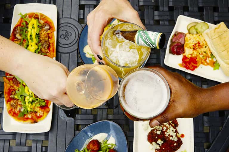 Overhead view of outdoor dining with pizzas, salads, and guests toasting with beverages on black patio table