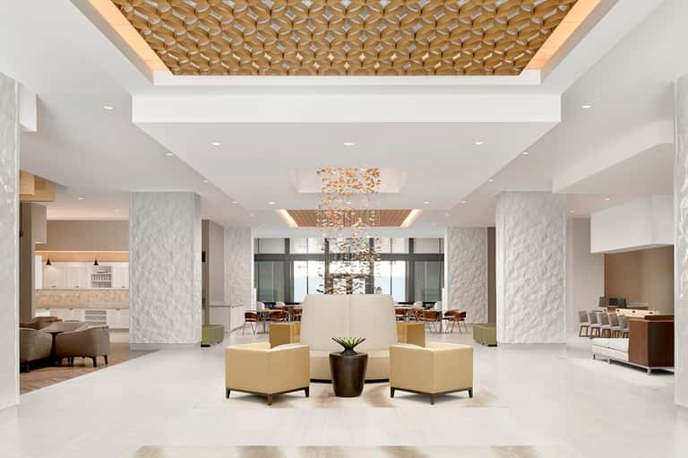 Bright hotel lobby featuring geometric wood ceiling pattern, marble columns, and beige seating areas