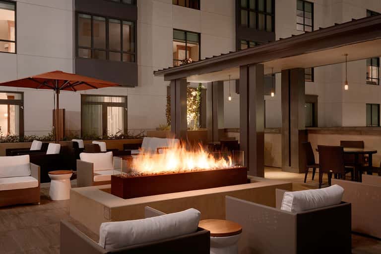Contemporary patio with fire pit feature wall, lounge seating, red umbrella, and modern building at dusk