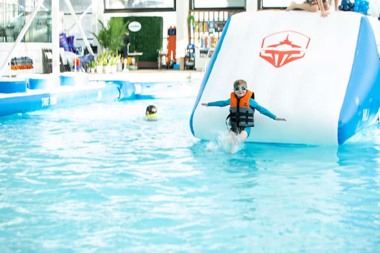 Child in life jacket splashing into indoor pool with inflatable obstacle course