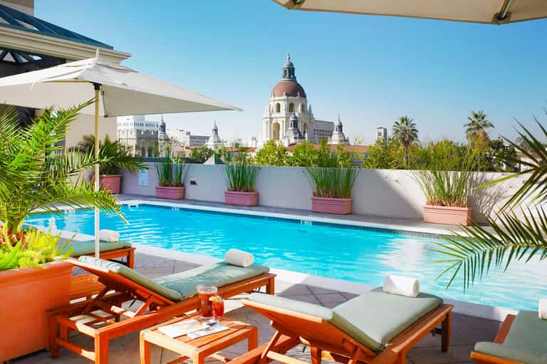 Rooftop pool with turquoise water, white loungers, palm plants, and cityscape with dome building in background